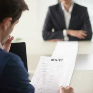 Back view of employer reading resume focus on document, job applicant waiting for hiring decision at desk on background. Hiring manager studying long list of candidates achievements and experiences