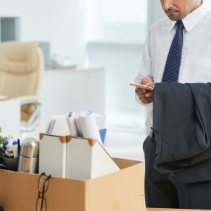 unrecognizable-man-standing-office-using-smartphone-with-personal-belongings-box